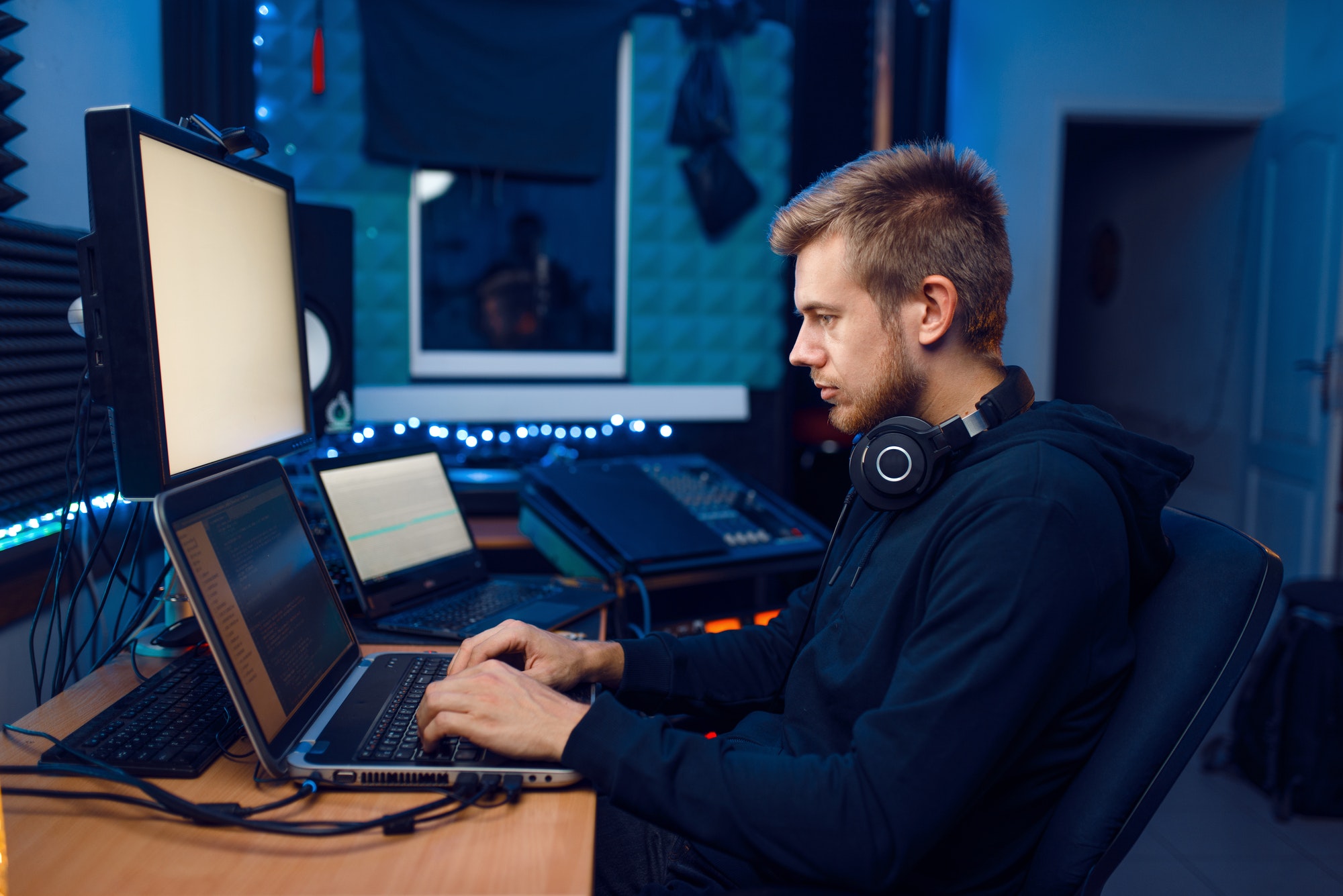 A man typing on a laptop with a second laptop and large monitor in a soundproofed studio
                        